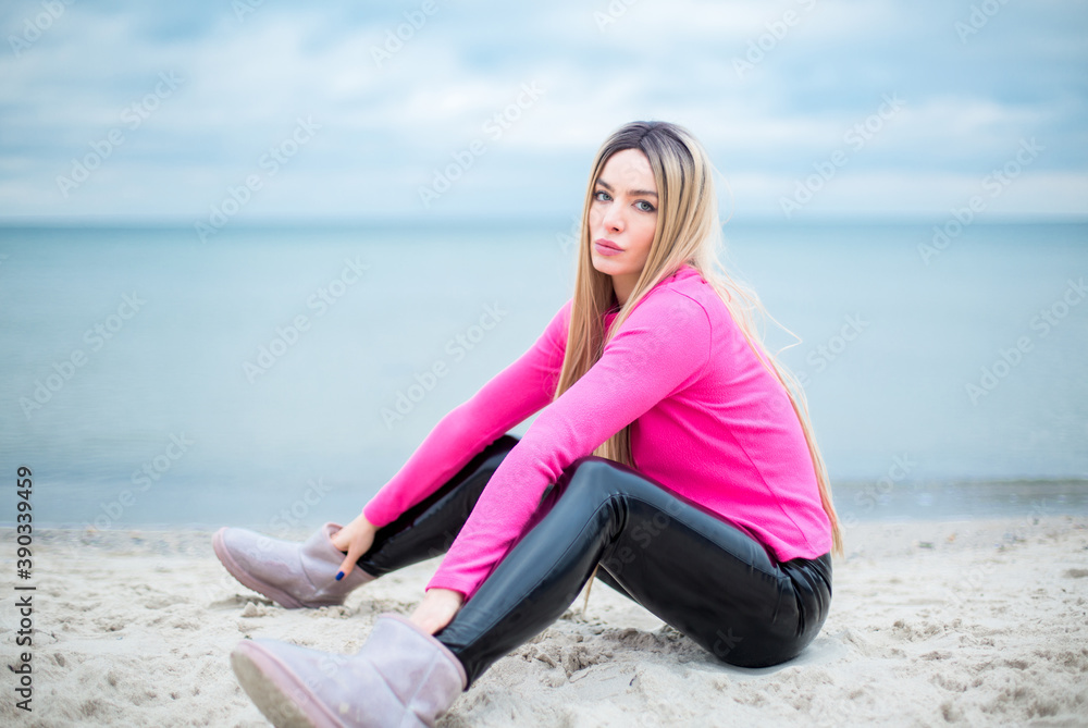 blonde caucasian female with blue eyes in patent leather trousers and crimson pullover is sitting on the sand at the beach, dramatic sky and sea at the background