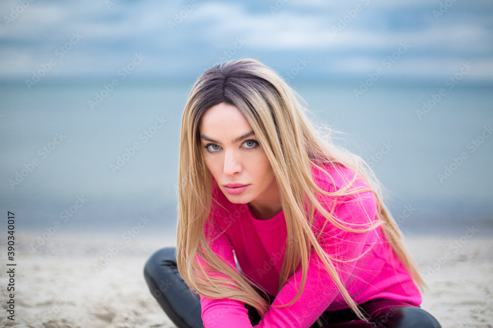 blonde caucasian female with blue eyes in patent leather trousers and crimson pullover is sitting on the sand at the beach, dramatic sky and sea at the background