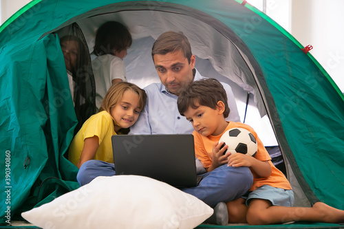 Caucasian dad sitting cross-legged with kids in tent at home and watching movie via laptop. Lovely children hugging father, having fun and playing. Childhood, family time and weekend concept