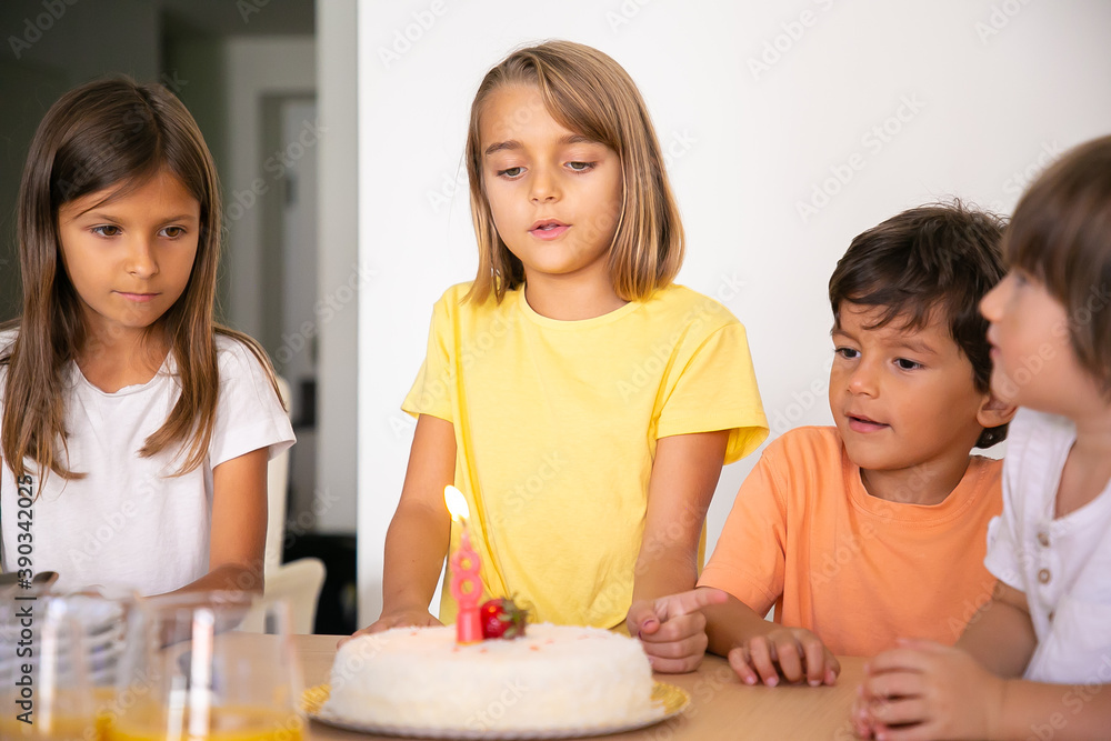Cute blonde girl making wish and celebrating her birthday with friends. Lovely children standing in room together and looking at tasty cake with candle. Childhood, celebration and holiday concept Cute blonde girl making wish and celebrating her birthday with friends. Lovely children standing in room together and looking at tasty cake with candle. Childhood, celebration and holiday concept