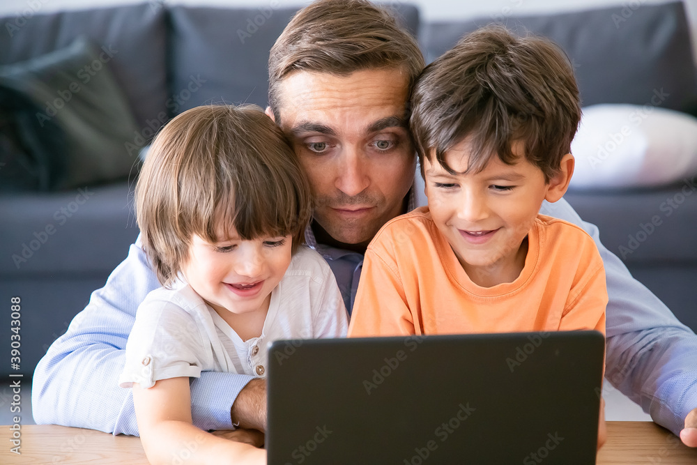 Middle-aged dad hugging sons and watching movie on laptop. Happy little boys sitting at table with father, looking at screen and smiling. Fatherhood, childhood and digital technology concept Middle-aged dad hugging sons and watching movie on laptop. Happy little boys sitting at table with father, looking at screen and smiling. Fatherhood, childhood and digital technology concept