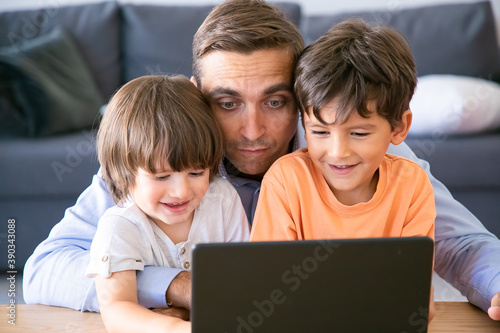 Middle-aged dad hugging sons and watching movie on laptop. Happy little boys sitting at table with father, looking at screen and smiling. Fatherhood, childhood and digital technology concept
