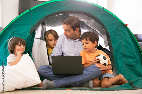 Thoughtful dad sitting cross-legged with kids in tent at home and holding laptop. Cute children watching movie on portable computer with Caucasian father. Childhood, family time and weekend concept