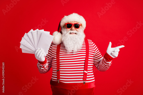 Photo of grandfather grey beard direct finger empty space hold fan envelopes wear santa claus x-mas costume suspenders sunglass striped shirt cap gloves isolated red color background © deagreez