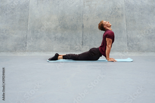 Fototapeta Naklejka Na Ścianę i Meble -  Young Man Stretching On Yoga Mat Against Concrete Wall Outdoors. Handsome Caucasian Sportsman With Strong Muscular Body In Fashion Sportswear Warming Up Before Intense Workout.