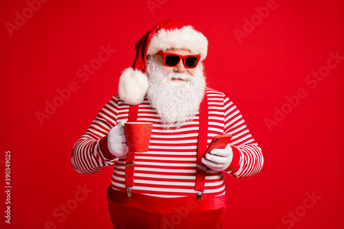 Portrait of his he nice handsome cheery focused bearded fat overweight Santa wear costume drinking caffeine using device 5g app isolated bright vivid shine vibrant red color background © deagreez
