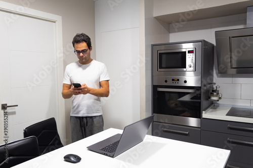 businessman working at home from kitchen, looking at mobile phone standing and his laptop on the table.