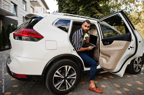 Successful arab man wear in striped shirt and sunglasses pose inside white suv car with laptop in hands.