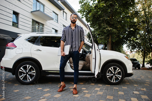 Successful arab man wear in striped shirt and sunglasses pose near his white suv car with laptop in hands. © AS Photo Family