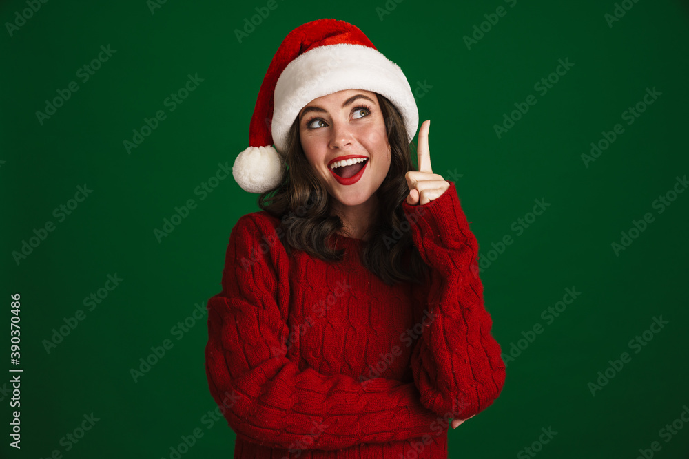 Excited girl in Santa Claus hat smiling and pointing finger upward Excited girl in Santa Claus hat smiling and pointing finger upward