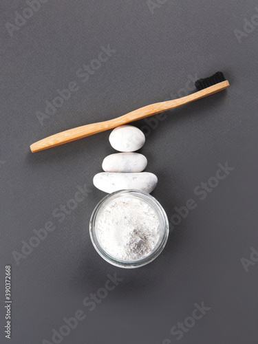 Zero waste concept for teeth cleaning. A bamboo toothbrush with the toothpowder in a glass decorating with three sea stones on the grey background.