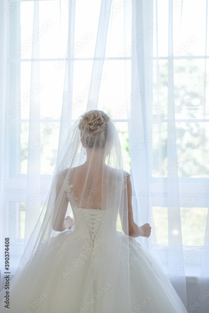 Bride in a apartment in a wedding dress. Bride looks out of the window, wedding day. Charming bride stand in front of window in a white wedding dress. Bride in a apartment in a wedding dress. Bride looks out of the window, wedding day. Charming bride stand in front of window in a white wedding dress.