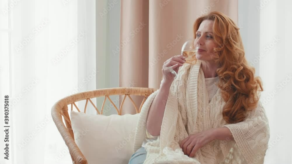Medium shot of attractive Caucasian middle-aged woman with long curly hair resting on rattan chair and drinking white wine Medium shot of attractive Caucasian middle-aged woman with long curly hair resting on rattan chair and drinking white wine