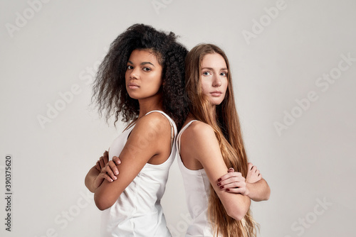 Portrait of two young diverse women wearing white shirts looking at camera while standing back to back isolated over grey background