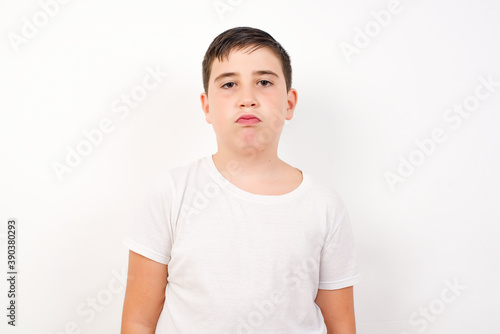 Joyful Caucasian young boy standing against white background looking to the camera, thinking about something. Both arms down, neutral facial expression.