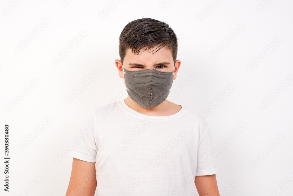 Caucasian young boy wearing medical mask standing against white background frowning his eyebrows being displeased with something. Caucasian young boy wearing medical mask standing against white background frowning his eyebrows being displeased with something.