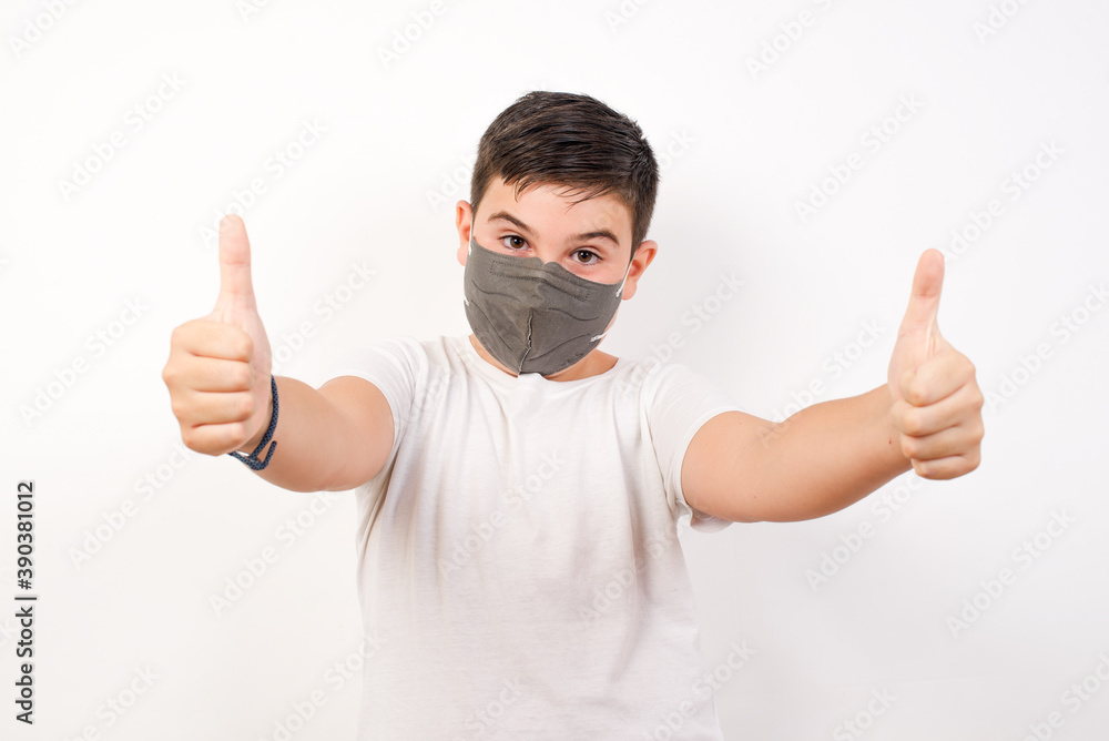 Portrait of happy smiling Caucasian young boy wearing medical mask standing against white background giving two thumbs up gesture: GOOD JOB! Positive human emotions. Portrait of happy smiling Caucasian young boy wearing medical mask standing against white background giving two thumbs up gesture: GOOD JOB! Positive human emotions.