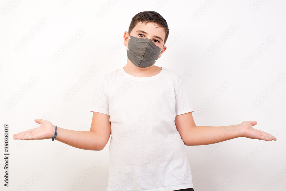 Puzzled and clueless Caucasian young boy wearing medical mask standing against white background with arms out, shrugging shoulders, saying: who cares, so what, I don't know. Negative human emotions. Puzzled and clueless Caucasian young boy wearing medical mask standing against white background with arms out, shrugging shoulders, saying: who cares, so what, I don't know. Negative human emotions.