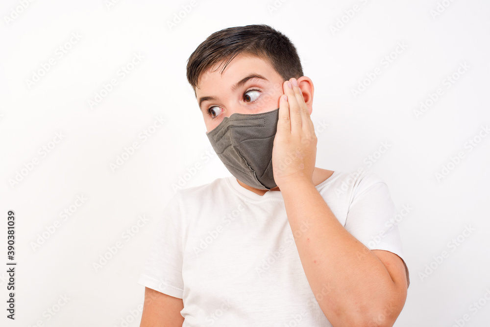 Caucasian young boy wearing medical mask standing against white background excited looking to the side hand on face. Advertisement and amazement concept. Caucasian young boy wearing medical mask standing against white background excited looking to the side hand on face. Advertisement and amazement concept.