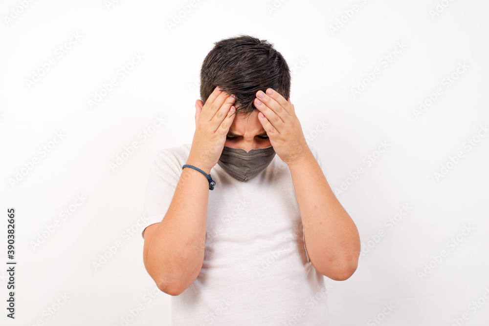 Caucasian young boy wearing medical mask standing against white background holding head with hands, suffering from severe headache, pressing fingers to temples Caucasian young boy wearing medical mask standing against white background holding head with hands, suffering from severe headache, pressing fingers to temples