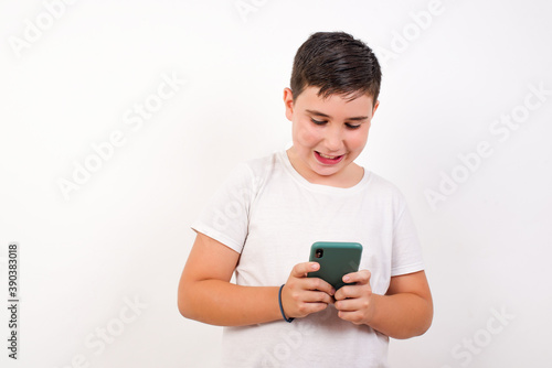 Smiling Caucasian young boy standing against white background using cell phone, messaging, being happy to text with friends, looking at smartphone. Modern technologies and communication. © Jihan