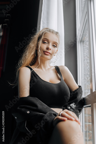 Stylish blonde in fashionable black clothes posing near the window.