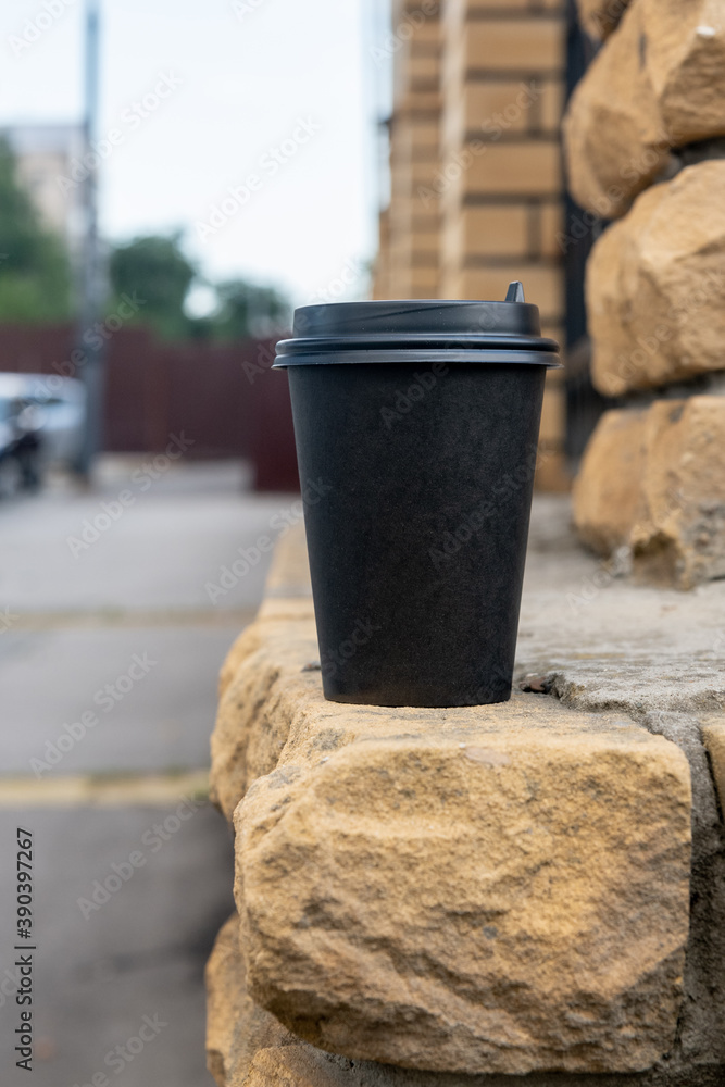 A black paper cup with coffee stands on a brick wall outside.