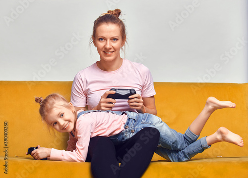 Excited young mother and cute little daughter sit on couch relax playing video games at home together, happy and small girls have fun in in living room.
