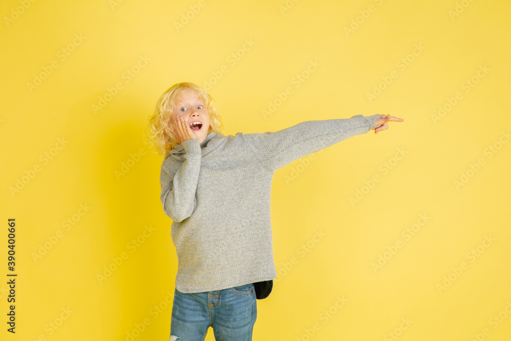 Pointing astonished. Portrait of little caucasian boy isolated on yellow studio background with copyspace. Beautiful model. Concept of human emotions, facial expression, sales, ad, youth. Pointing astonished. Portrait of little caucasian boy isolated on yellow studio background with copyspace. Beautiful model. Concept of human emotions, facial expression, sales, ad, youth.