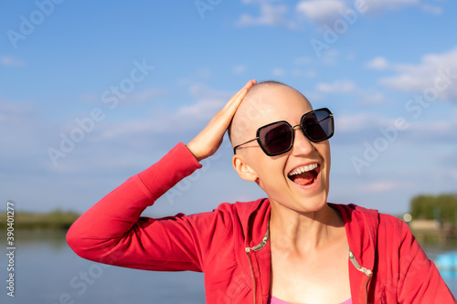 Happy young caucasian bald confident hipster woman shoving shaved head enjoying life after surviving breast cancer. Portrait of beautiful hairless girl smiling walking near lake at bright sunny day