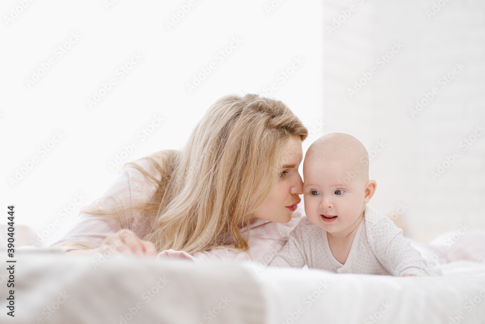 Young beautiful Caucasian blonde mom lies on the bed with her baby daughter. White light home interior. Young beautiful Caucasian blonde mom lies on the bed with her baby daughter. White light home interior.