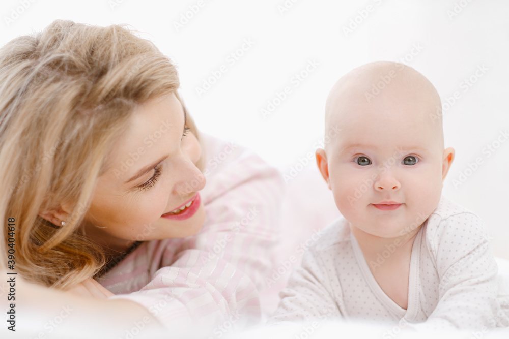 Young beautiful Caucasian blonde mom lies on the bed with her baby daughter. White light home interior. Young beautiful Caucasian blonde mom lies on the bed with her baby daughter. White light home interior.