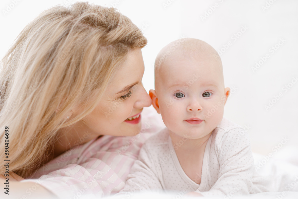 Young beautiful Caucasian blonde mom lies on the bed with her baby daughter. White light home interior. Young beautiful Caucasian blonde mom lies on the bed with her baby daughter. White light home interior.