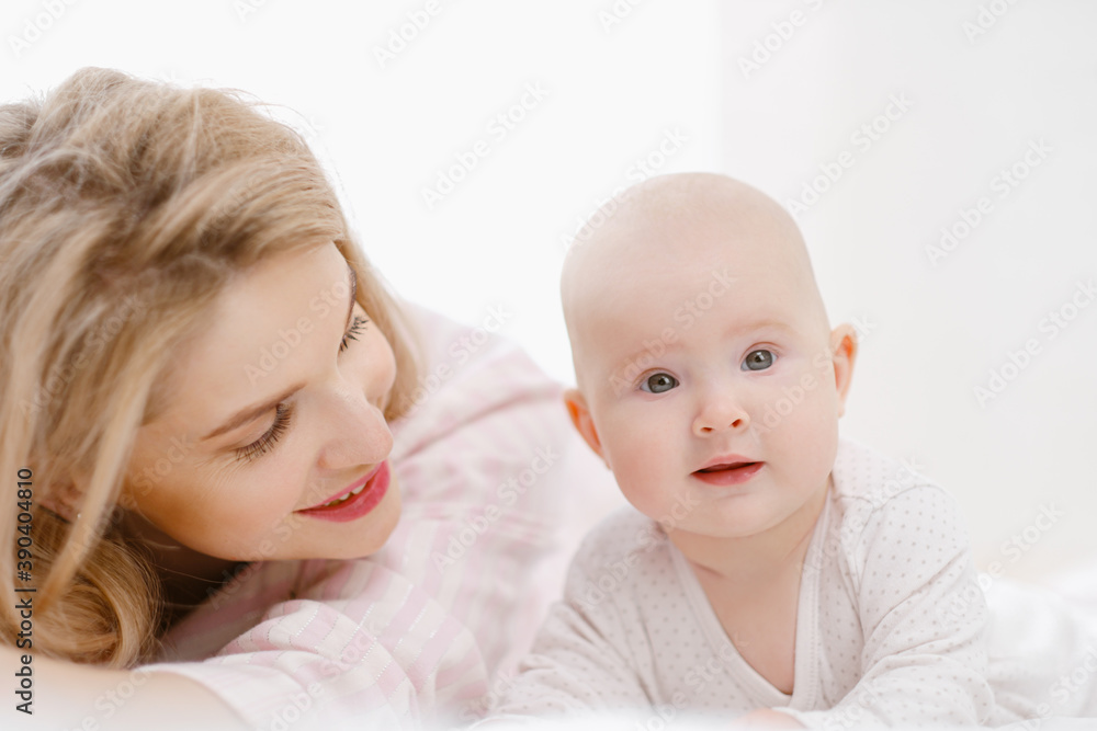 Young beautiful Caucasian blonde mom lies on the bed with her baby daughter. White light home interior. Young beautiful Caucasian blonde mom lies on the bed with her baby daughter. White light home interior.