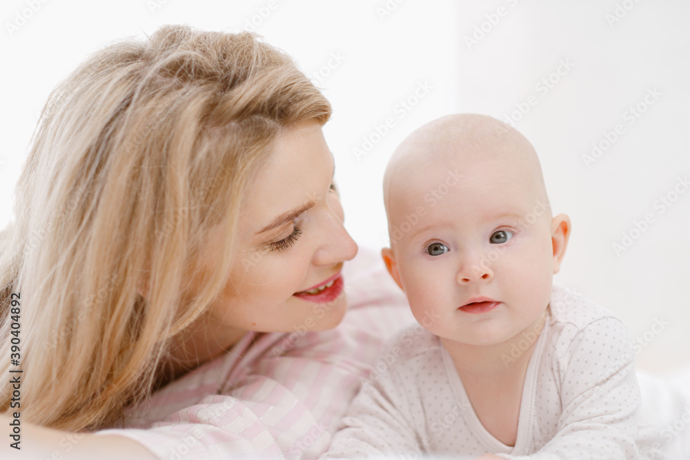Young beautiful Caucasian blonde mom lies on the bed with her baby daughter. White light home interior. Young beautiful Caucasian blonde mom lies on the bed with her baby daughter. White light home interior.