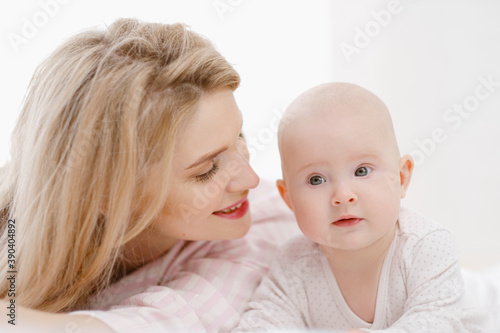 Young beautiful Caucasian blonde mom lies on the bed with her baby daughter. White light home interior.