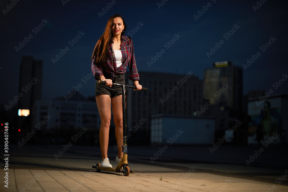Beautiful sexy young girl in short shorts posing on scooter  in city in evening.