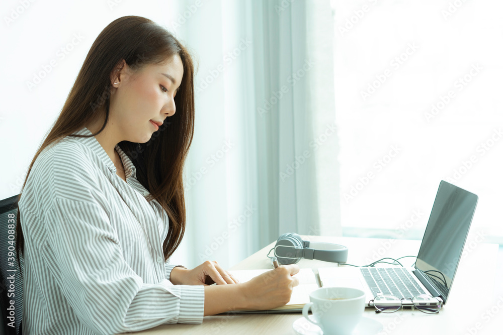 portrait a beautiful young woman taking notes and learning online with a laptop sitting in a table in the living room at the home office, work from home concept. portrait a beautiful young woman taking notes and learning online with a laptop sitting in a table in the living room at the home office, work from home concept.