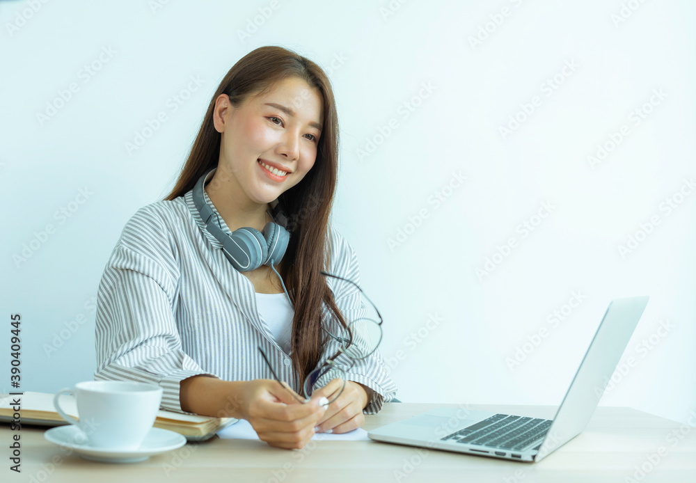 portrait of a beautiful Asian woman wearing headphones Taking off his glasses and smiling at the camera happily. On her desk At the home office, the idea of working at home. portrait of a beautiful Asian woman wearing headphones Taking off his glasses and smiling at the camera happily. On her desk At the home office, the idea of working at home.