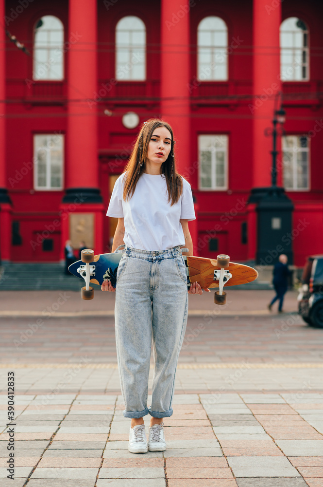 Fototapeta premium Stylish female skater in stylish casual clothes stands on the street and poses with a gongboard behind her back, looking at the camera with a serious face. Vertical