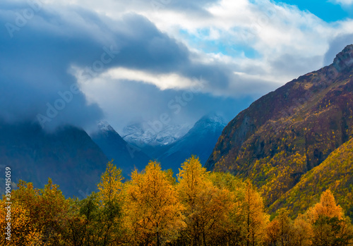 Valley with trees in autumn colors , dramatic cloud scape and mountain slopes and peaks in the distance.