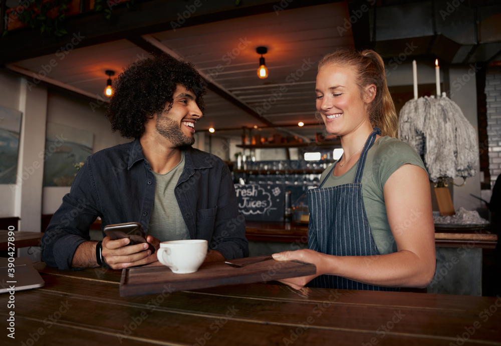 Cheerful female waitress serving coffee to young male customer with curly hair using smartphone in modern cafe