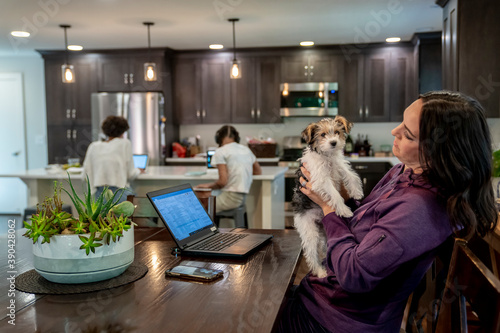 Woman working from home on laptop computer while holding cute puppy, daughters study at kitchen counter in background