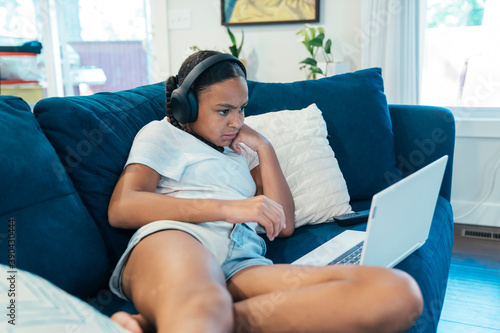 Girl in wireless headphones sitting in living room doing home school work on laptop