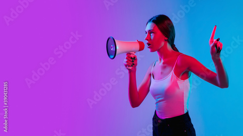 Pointing with loudspeaker. Young caucasian woman's portrait on gradient blue-purple studio background in neon. Concept of youth, human emotions, facial expression, sales, ad. Beautiful model. Flyer