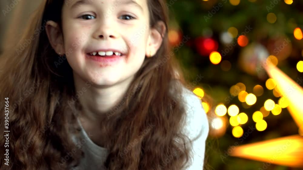 Portrait of happy little girl sitting near Christmas tree on new year's eve in decorated room. New Year and holidays with kids concept. Enjoying festive holiday celebration. 4k video Portrait of happy little girl sitting near Christmas tree on new year's eve in decorated room. New Year and holidays with kids concept. Enjoying festive holiday celebration. 4k video