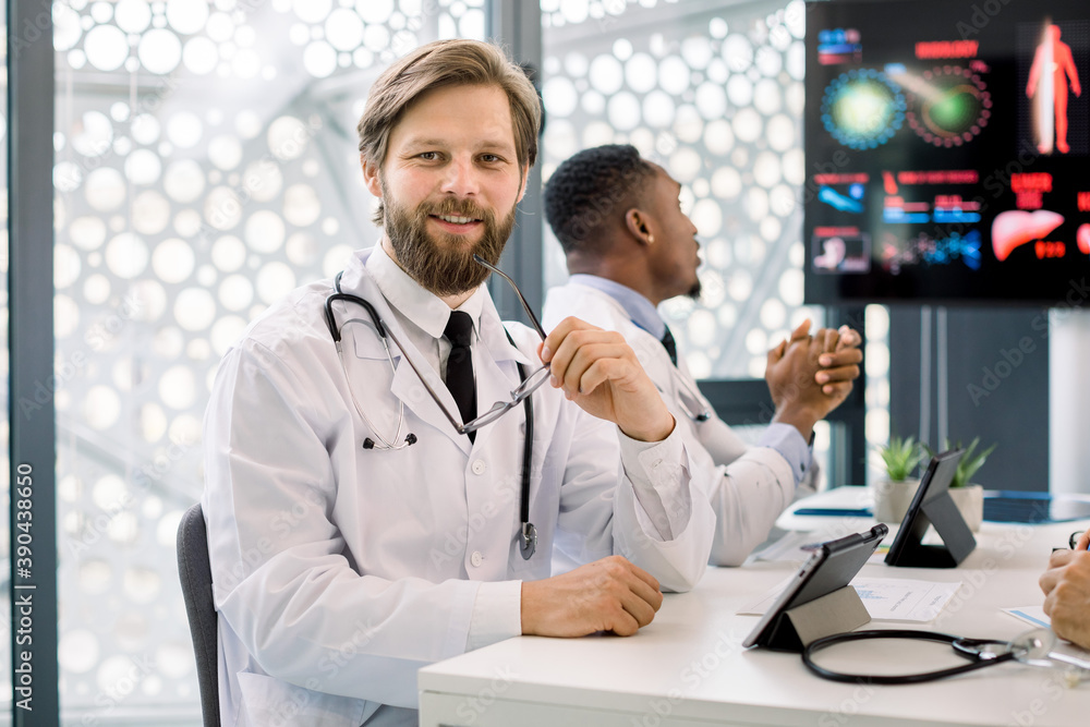 Portrait of handsome smiling young Caucasian bearded man doctor, sitting at the table during medical staff meeting or seminar and using tablet computer. African male doctor on the background Portrait of handsome smiling young Caucasian bearded man doctor, sitting at the table during medical staff meeting or seminar and using tablet computer. African male doctor on the background