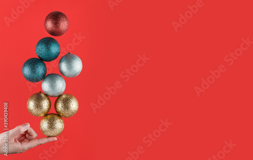 Person holding with one finger brightly colored Christmas spheres striking a balance on a red background with copy space. Christmas decorations. Christmas wallpaper