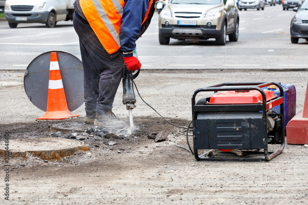 A worker repairs a section of the road with an electric jackhammer against the background of passing cars in blur.