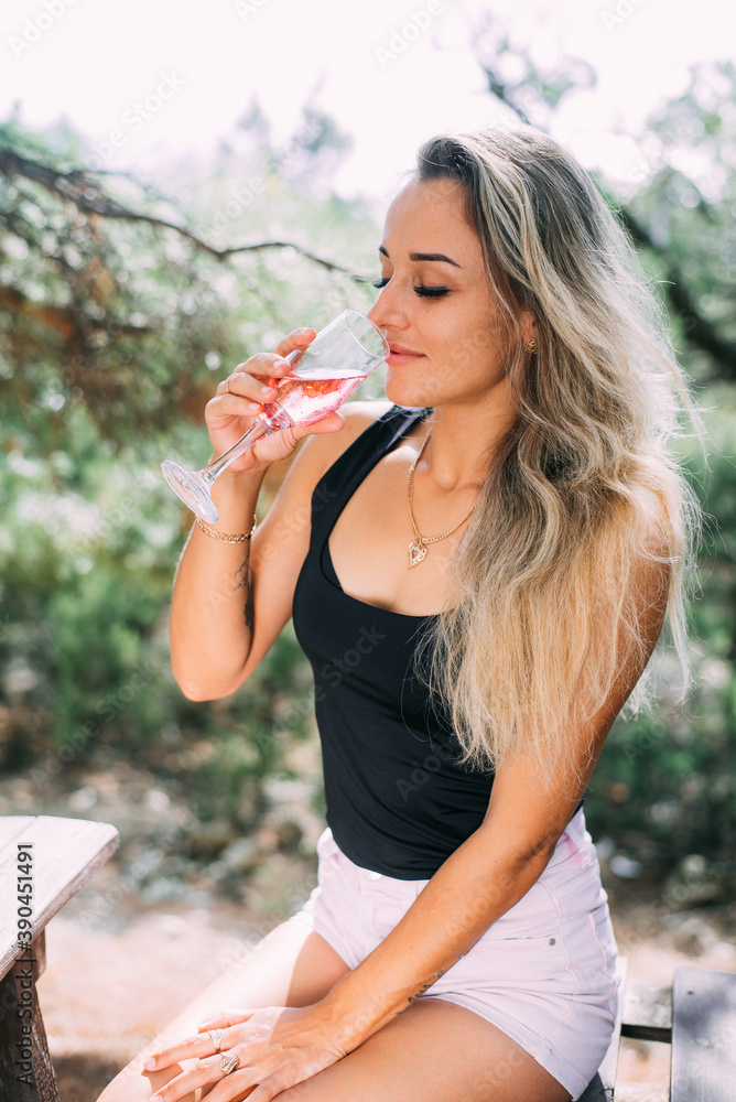 Fototapeta premium Beautiful girl in a juniper and pine forest drinking wine or champagne sitting on a wooden pallet, during the day
